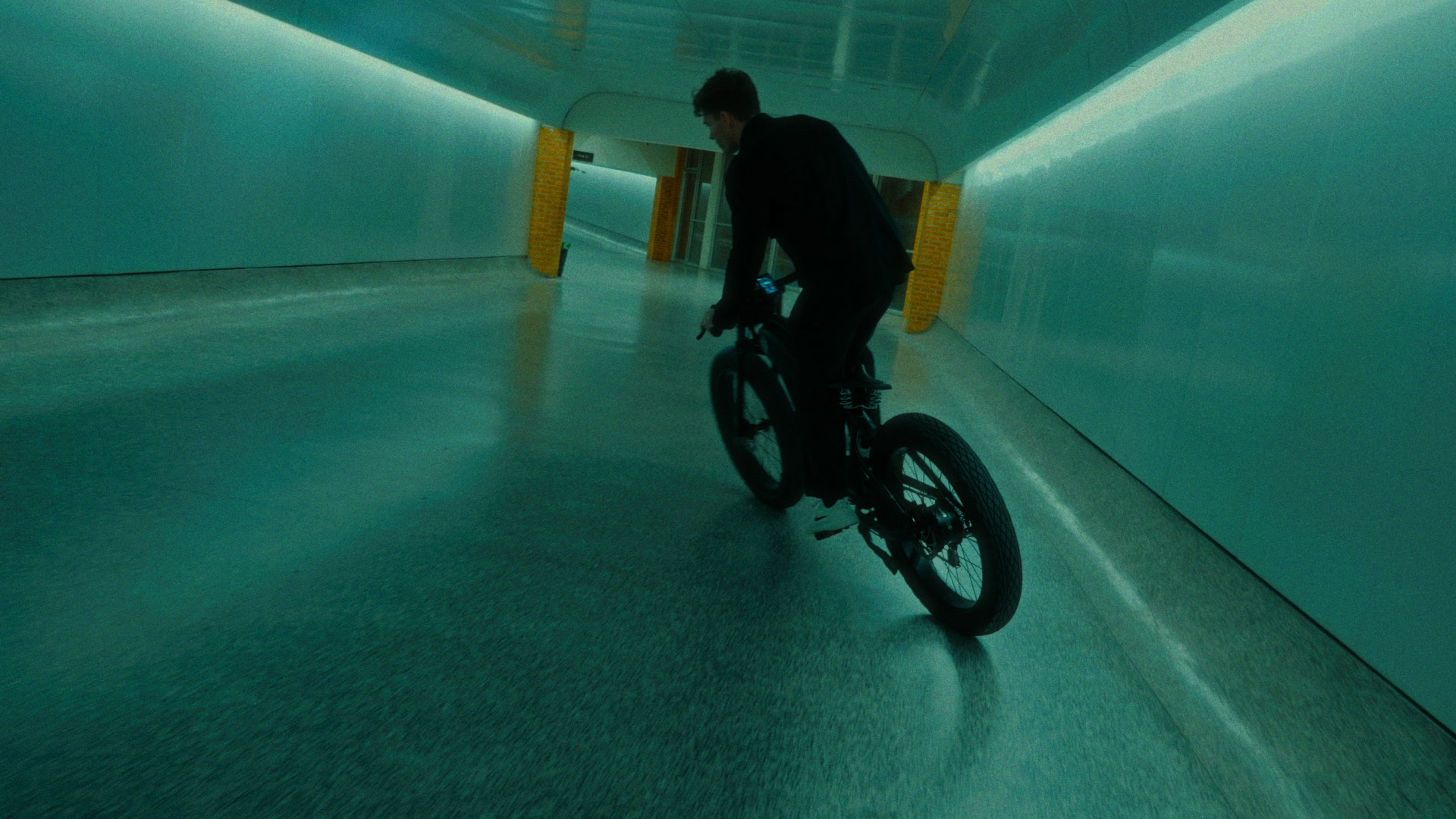 Wide teal tunnel frame with a rider moving through polished concrete and practical strip light.