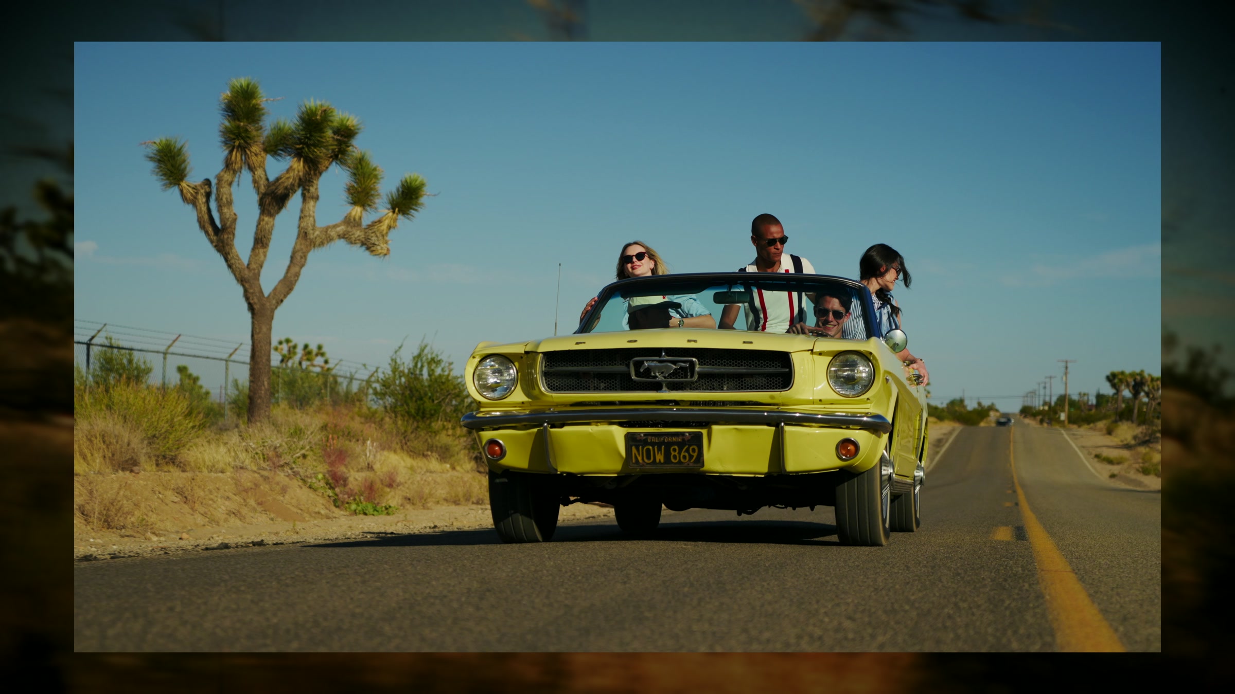 Yellow convertible moving through open desert road with fashion styling, hard sun, and mountain horizon.