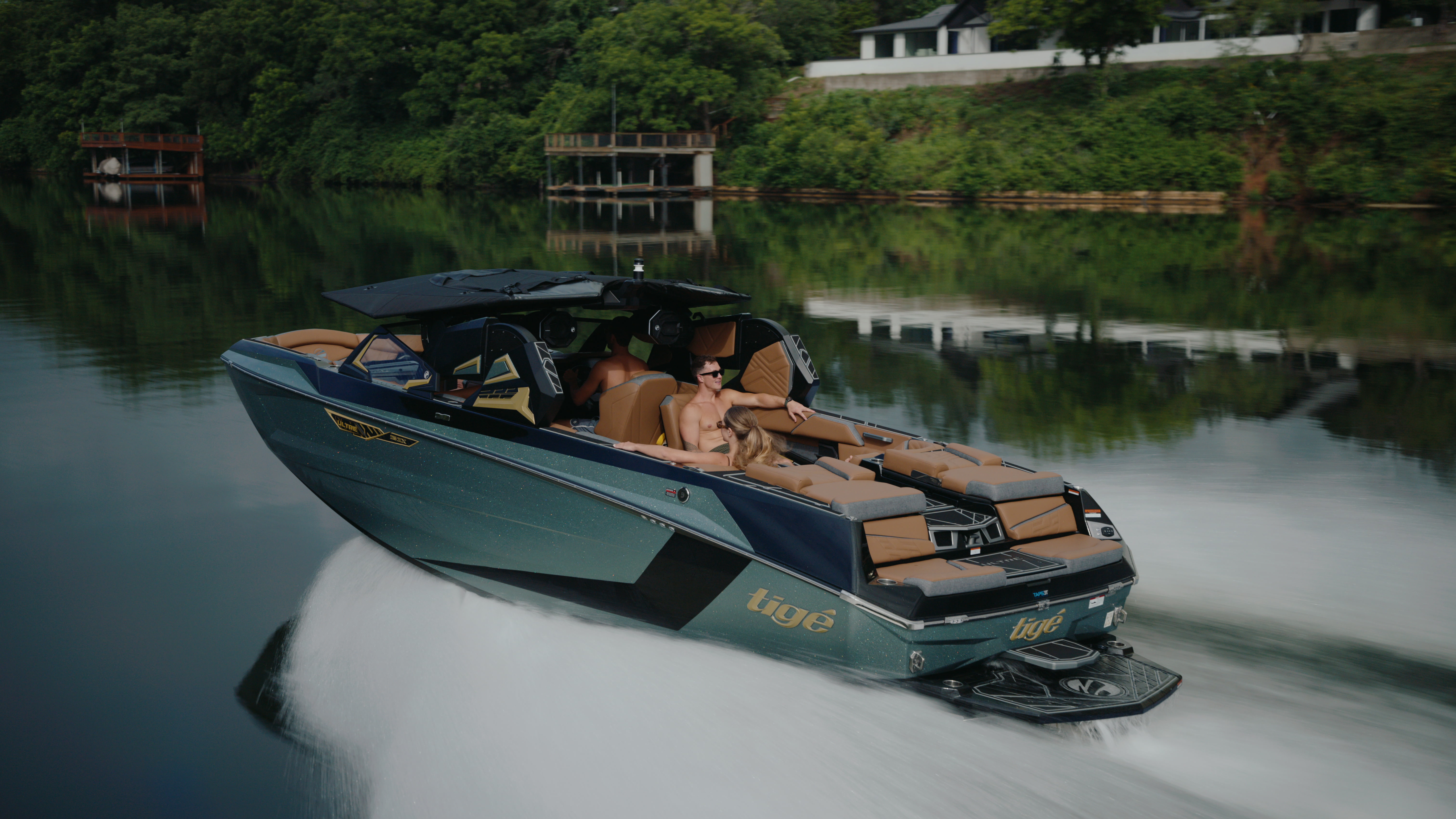 Cinematic lakeside pass of a Tige boat with passengers, a glassy wake, and wooded shoreline reflections.